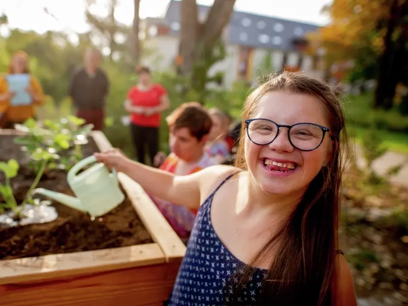 Eine junge Frau mit Down-Syndrom gießt Pflanzen in einem Hochbeet. Sie lacht fröhlich direkt in die Kamera.