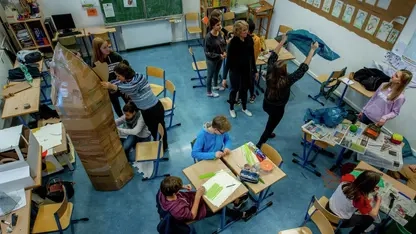 Ein Klassenzimmer von oben. Einige Kinder sitzen an einem Tisch, andere stehen. Es wird gebastelt.