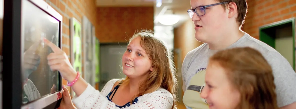 Zwei Mädchen und ein Junge stehen in einem Schulflur und schauen auf einen Monitor. Ein Mädchen zeigt auf den Monitor.