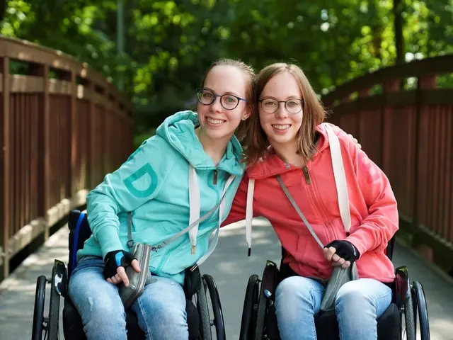 Leona und Ellen lachen in die Kamera. Sie sitzen beide Rollstuhl.