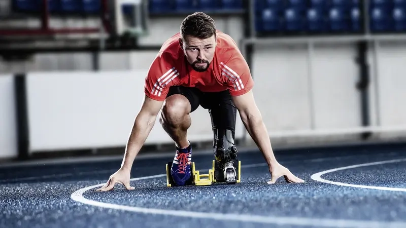 Junger Mann mit dunklen Haaren und kurzem Vollbart kniet in einem Startblock auf einer Laufbahn
