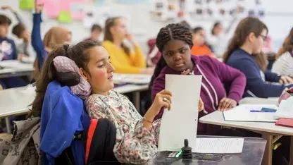 Ein junges Mädchen im Rollstuhl sitz an einem Tisch und hat ein Papier in der Hand. Ihre Banknachbarin schaut auch auf das Blatt Papier.