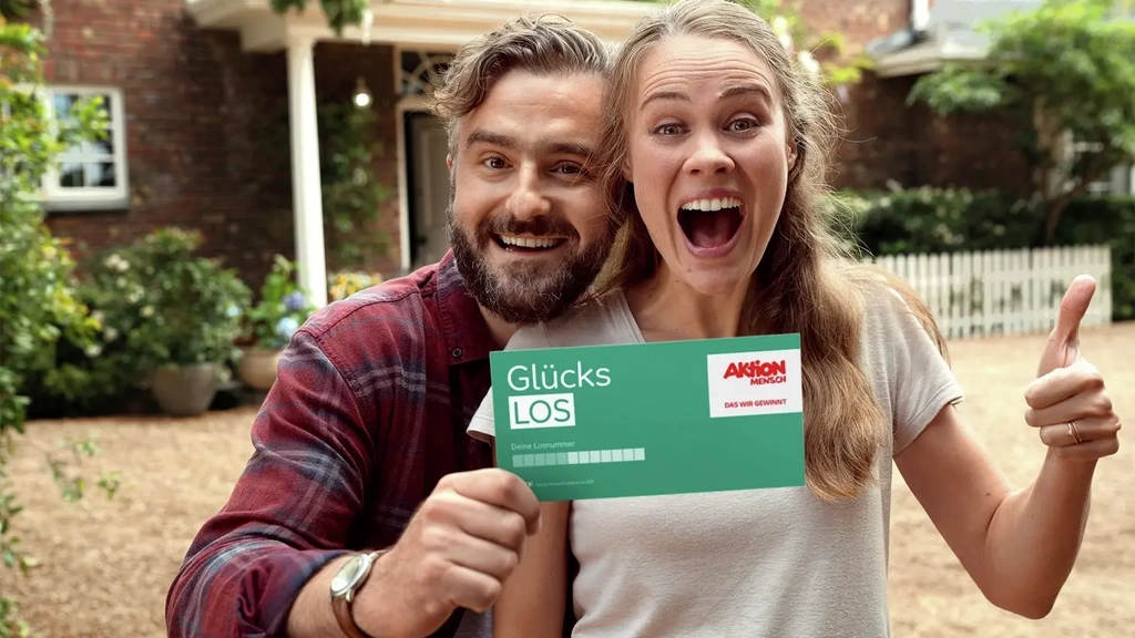 Ein Mann und eine Frau legen die Köpfe aneinander und lachen jubelnd. Sie halten ein Glückslos in die Kamera.