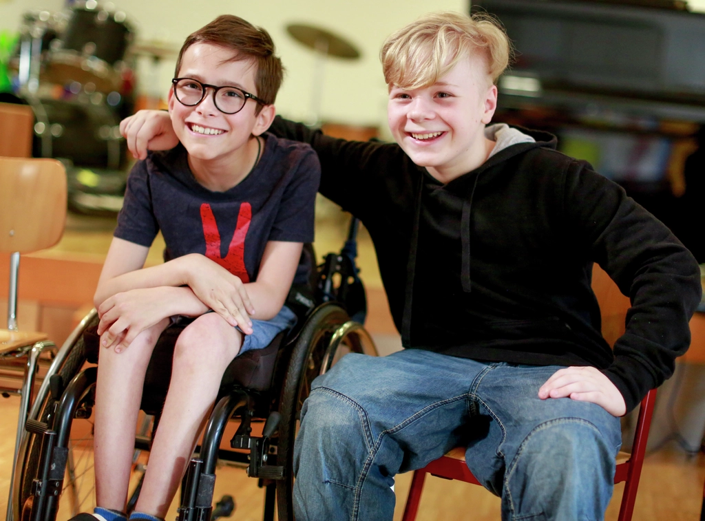 Zwei Jungen sitzen nebeneinander. Der eine auf einem Stuhl, der andere in einem Rollstuhl. Der Junge auf dem Stuhl hat den Arm um den Jungen im Rollstuhl gelegt. Beide lachen in die Kamera.
