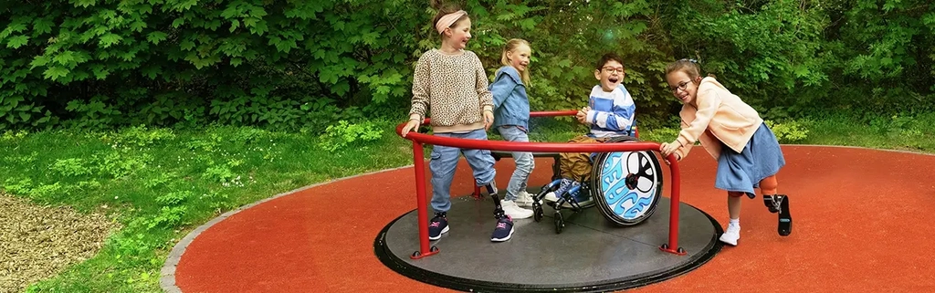Ein Mädchen mit Beinprothese schiebt ein Inklusions-Karussell an. Auf dem Karussell befinden sich ein Junge, der im Rollstuhl sitzt, und zwei Mädchen.