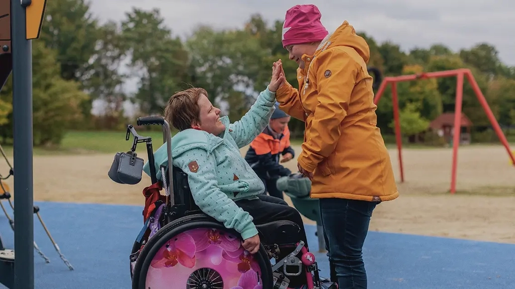 Zwei Mädchen klatschen sich mit der Hand ab. Ein Mädchen ist Rollifahrerin.