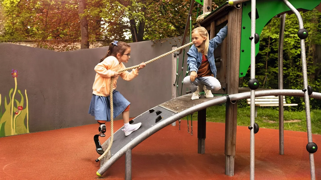 Zwei Mädchen spielen auf einem barrierefreien Spielturm. Ein Mädchen mit Beinprothese erklimmt den Turm gerade, indem sie sich an einem Seil über eine Rampe hochzieht.