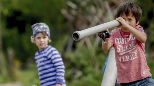 Zwei Kinder spielen. Ein Junge schaut durch ein schmales Rohr, als suche er etwas.