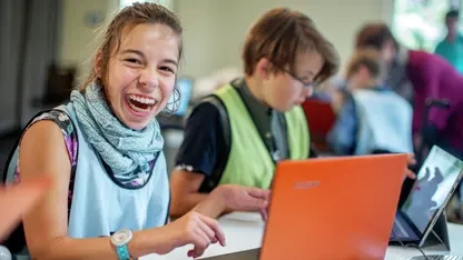 Ein Mädchen sitzt an einem Laptop und lächelt in die Kamera. Neben ihr sitzt ein Junge an einem Laptop.