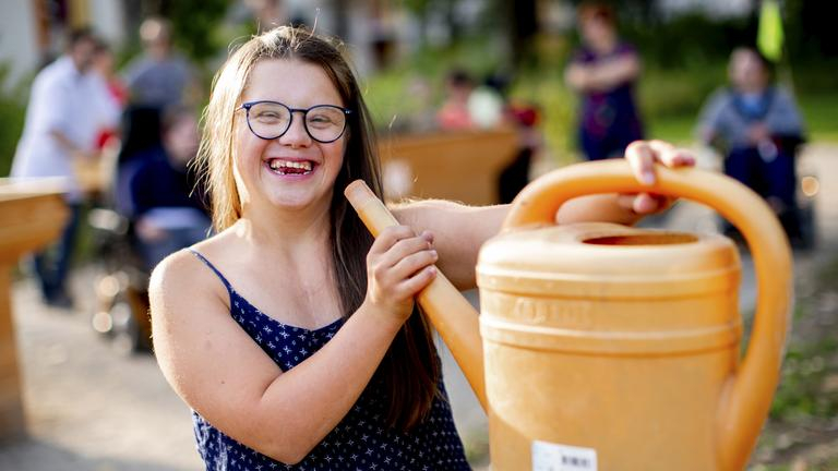 Eine junge Frau mit Down-Syndrom hält eine Gießkanne im Spaß wie ein Megaphon an ihren Mund. Sie lacht fröhlich in die Kamera.