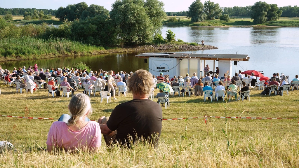 Das Traumschüff liegt  in Dömitz vor Anker. Am Ufer sitzt das Publikum und verfolgt die Theatervorstellung.